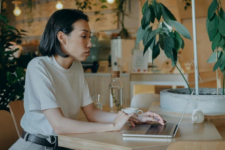 Asian woman working on a laptop in a cozy coffee shop setting, surrounded by plants.